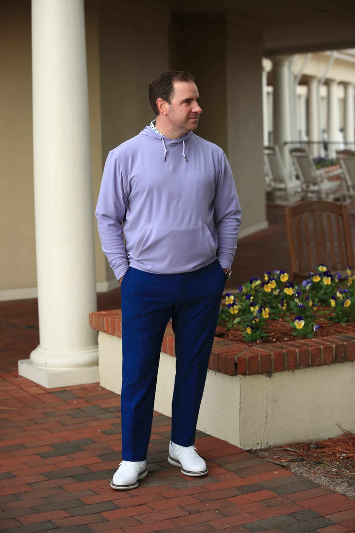 Man in a light purple hoodie and blue pants stands by a flowerbed with yellow and purple flowers, looking away