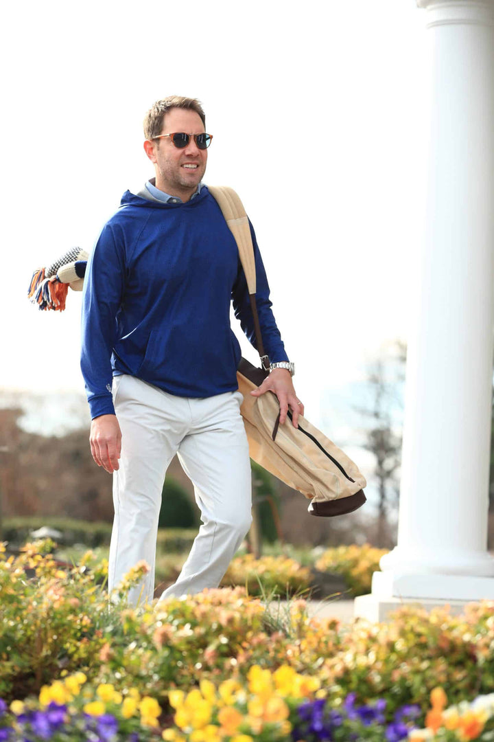 Man in a blue hoodie and white pants walks happily with a beige golf bag over his shoulder