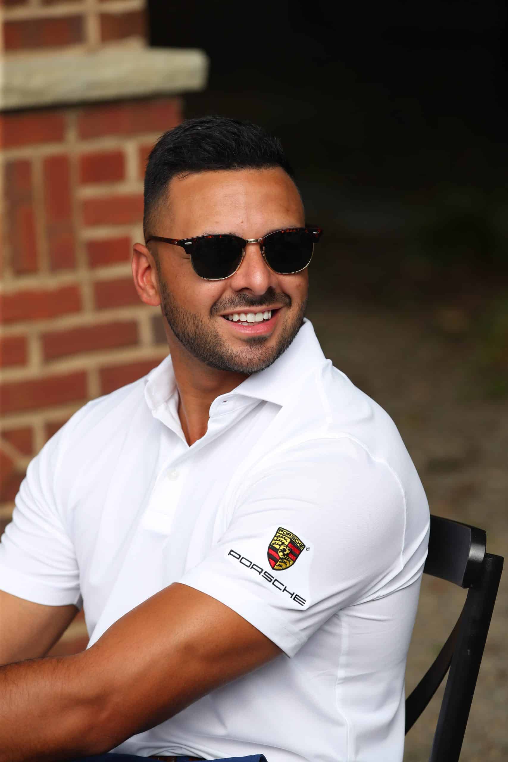 A man in a white Porsche polo shirt and sunglasses smiles while seated