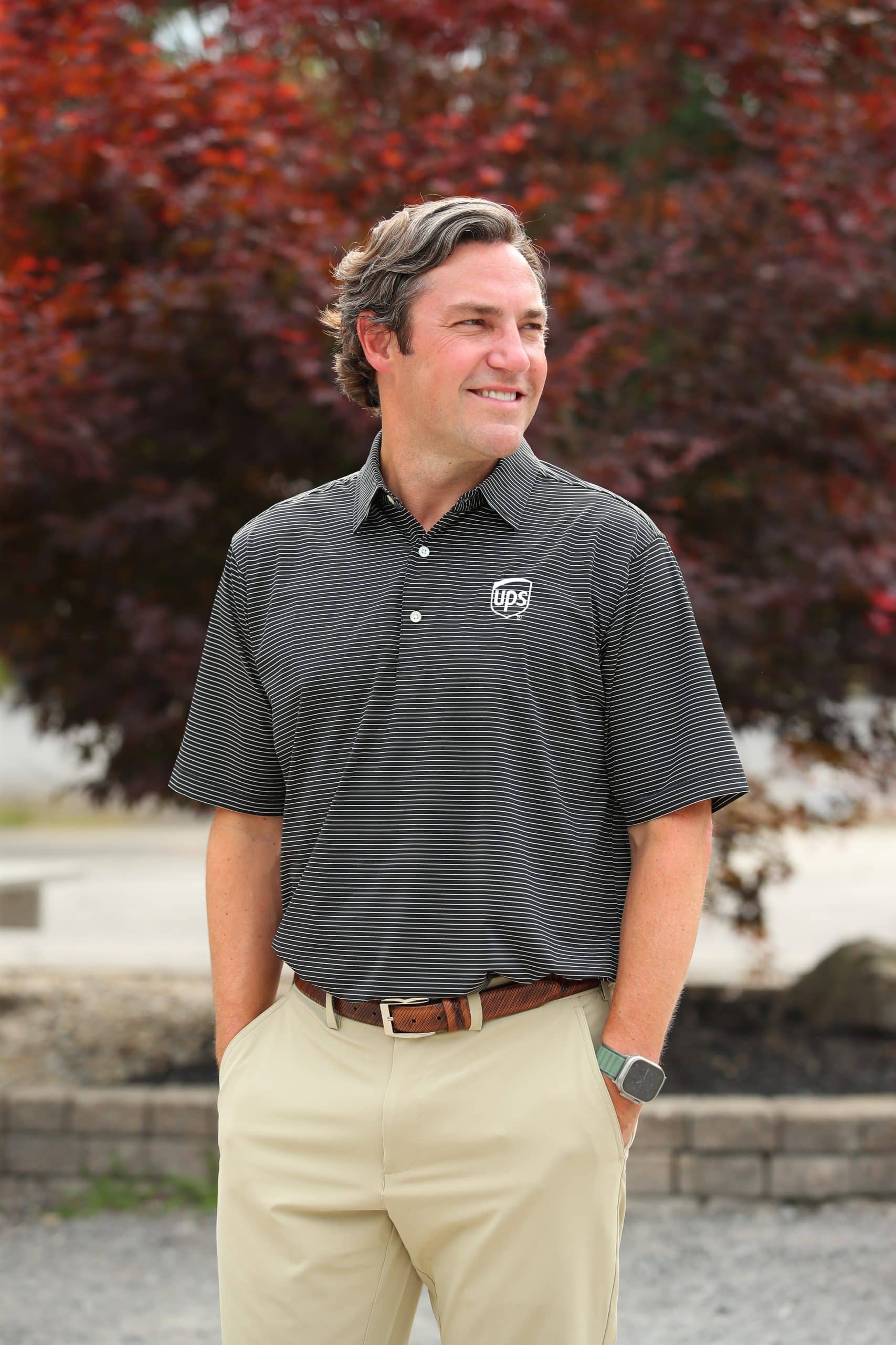Man in a striped UPS polo and beige pants, stands outdoors with hands in pockets, smiling and looking to the right