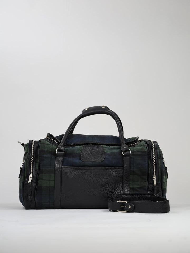 A duffel bag with a green and blue plaid fabric is pictured against a plain background. It features black leather handles and a detachable strap, with silver zippers