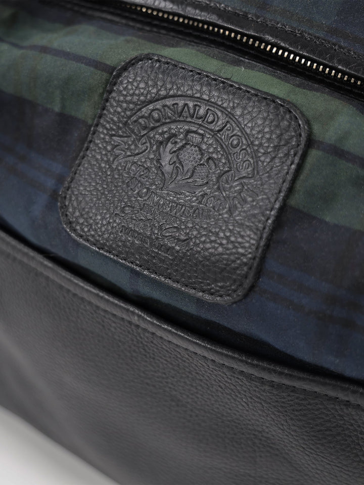 Close-up of a black leather bag with embossed logo on a textured patch, set against a green and blue plaid fabric lining