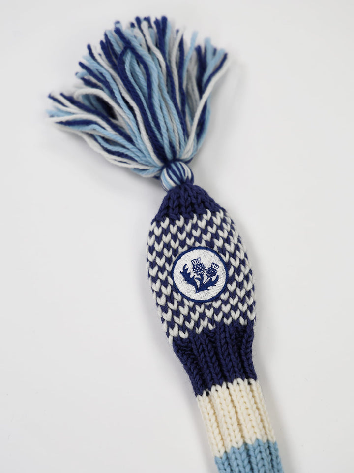 A knit golf club headcover with blue, white, and navy patterns features a fringe top and a thistle emblem on the front