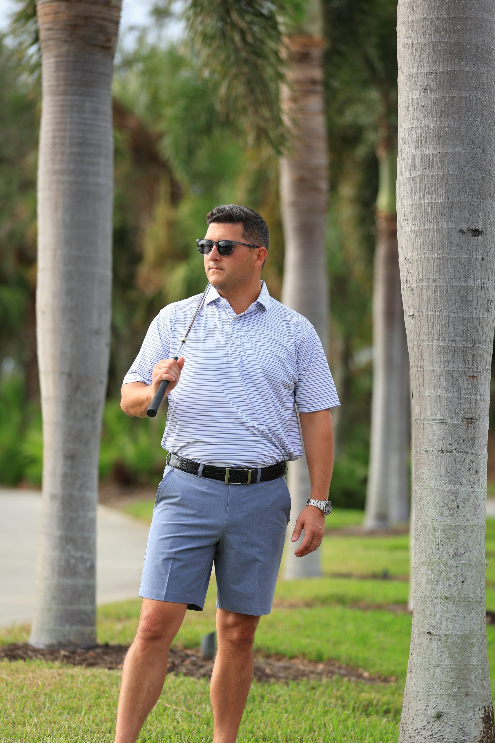 A man in sunglasses, wearing a striped polo and blue shorts, holds a golf club while standing among tall palm trees