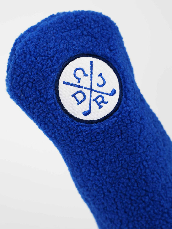 Close-up of a fluffy blue golf club headcover. It features a circular white patch with blue crossed golf clubs and letters.