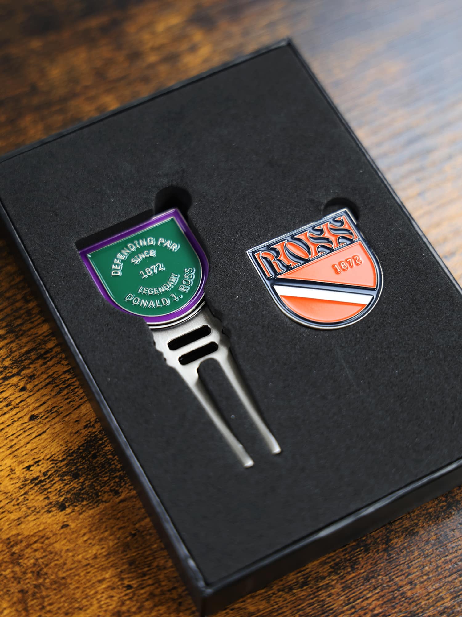 A black box holds a golf divot tool and ball marker. The tool has a green, purple, and silver shield with text, while the marker is orange
