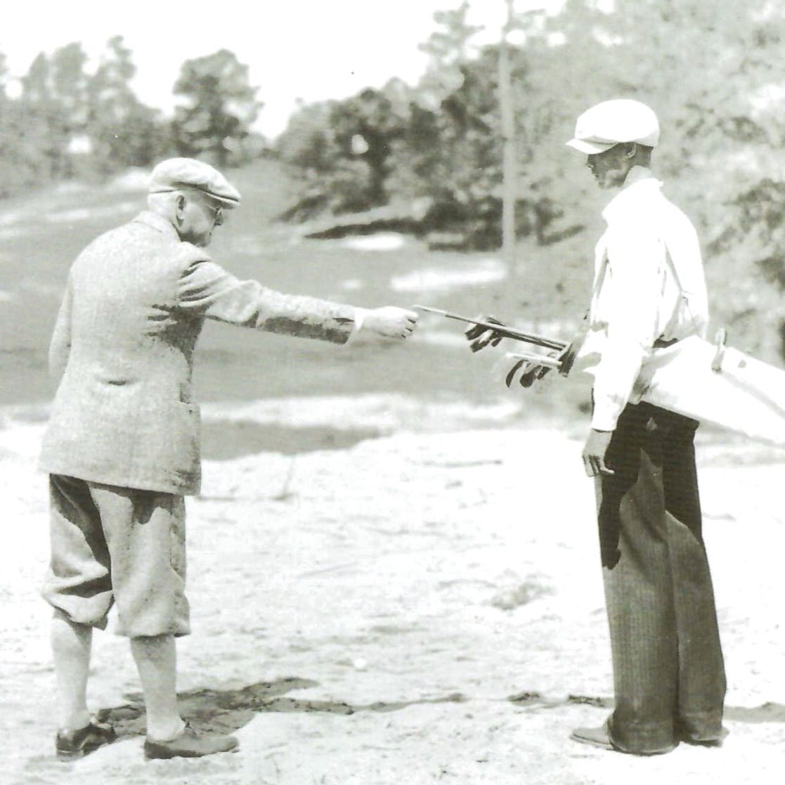 Vintage photo of two men on a golf course. One, in knickers and cap, exchanges a club with a caddie holding a bag, set against a blurred tree backdrop