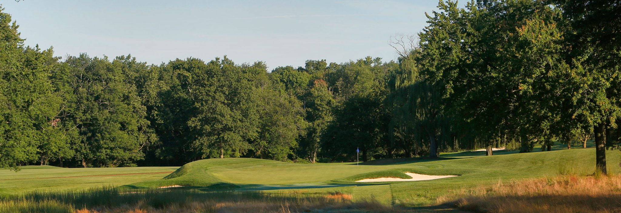 Lush golf course with a well-maintained green, sand bunkers, and a flagpole. Course is surrounded by dense trees under a clear blue sky