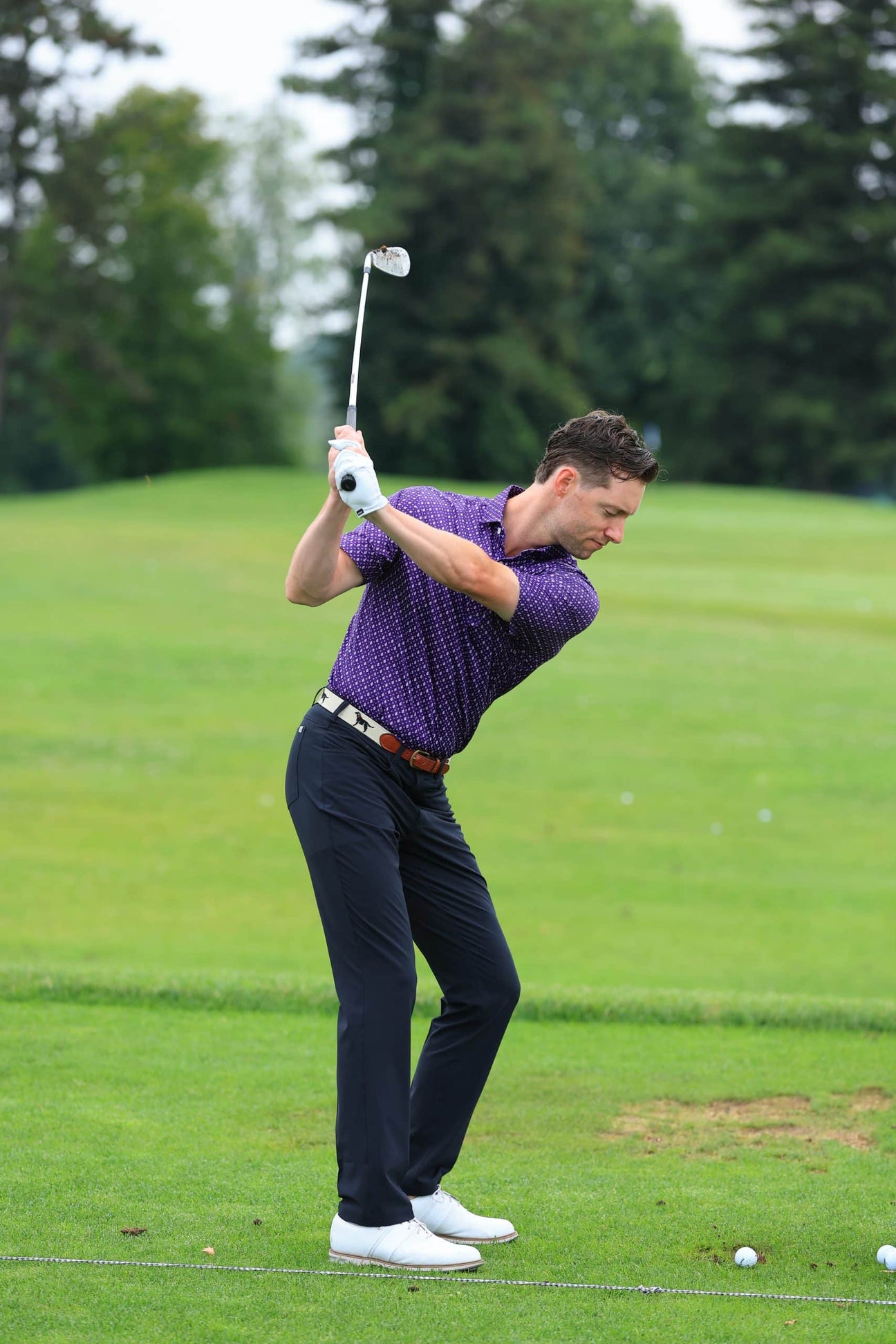 A man in a purple polo and dark pants swings a golf club on a lush green course. His focused expression conveys concentration and athleticism.