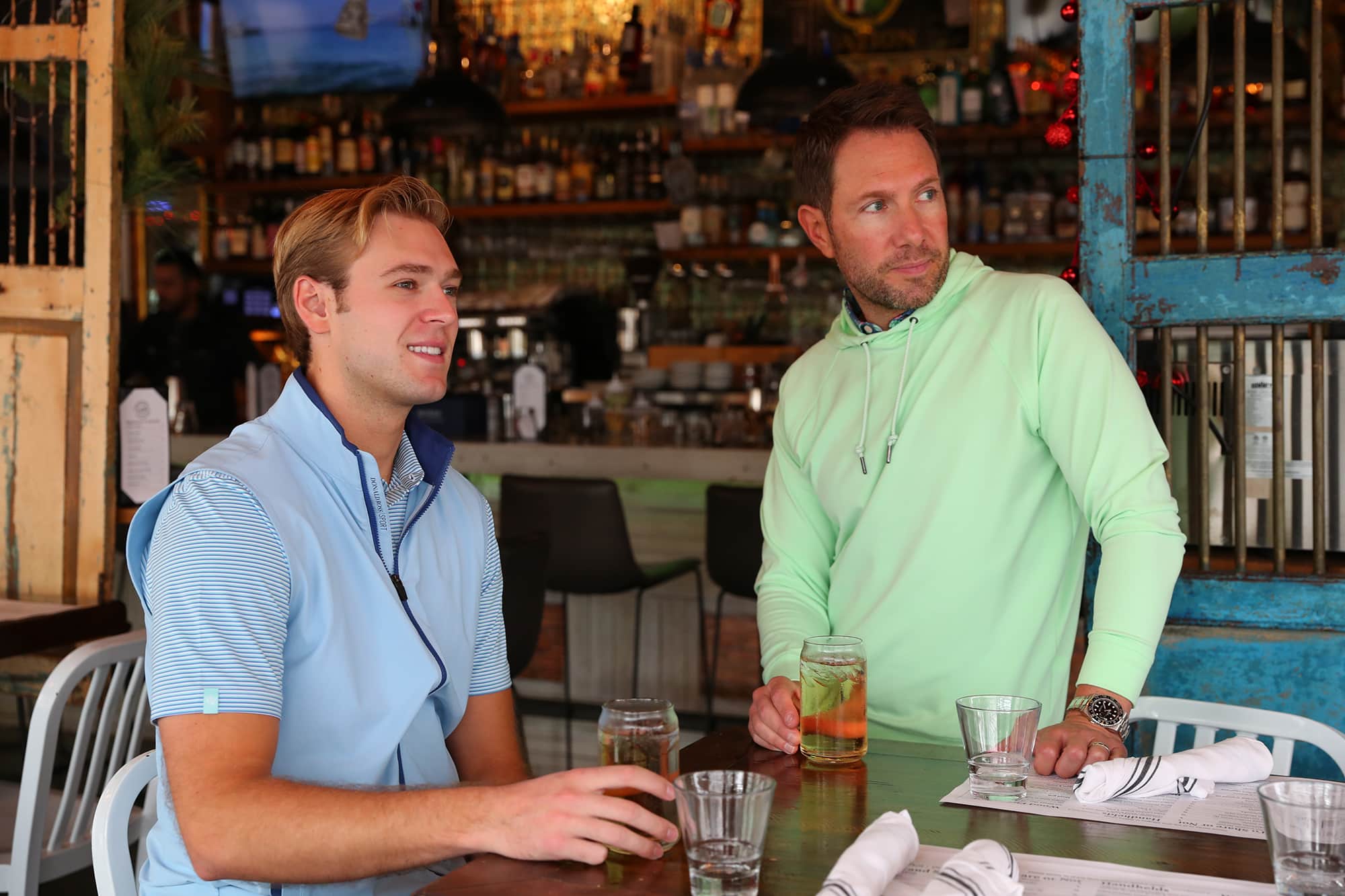 Two men are sitting at a table in a bar, engaging in conversation while holding drinks.