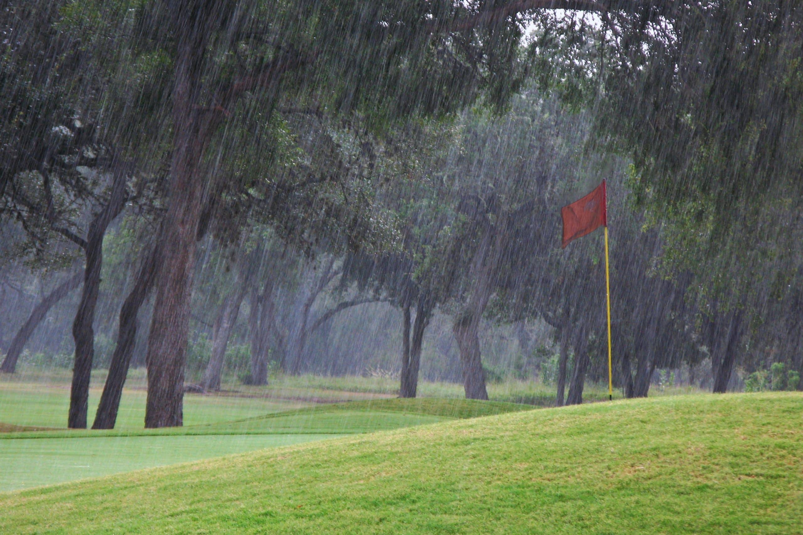 A golf course is seen in heavy rain, with a lush green landscape. A red flag on a yellow pole stands prominently, surrounded by tall trees. The mood is serene yet dramatic.