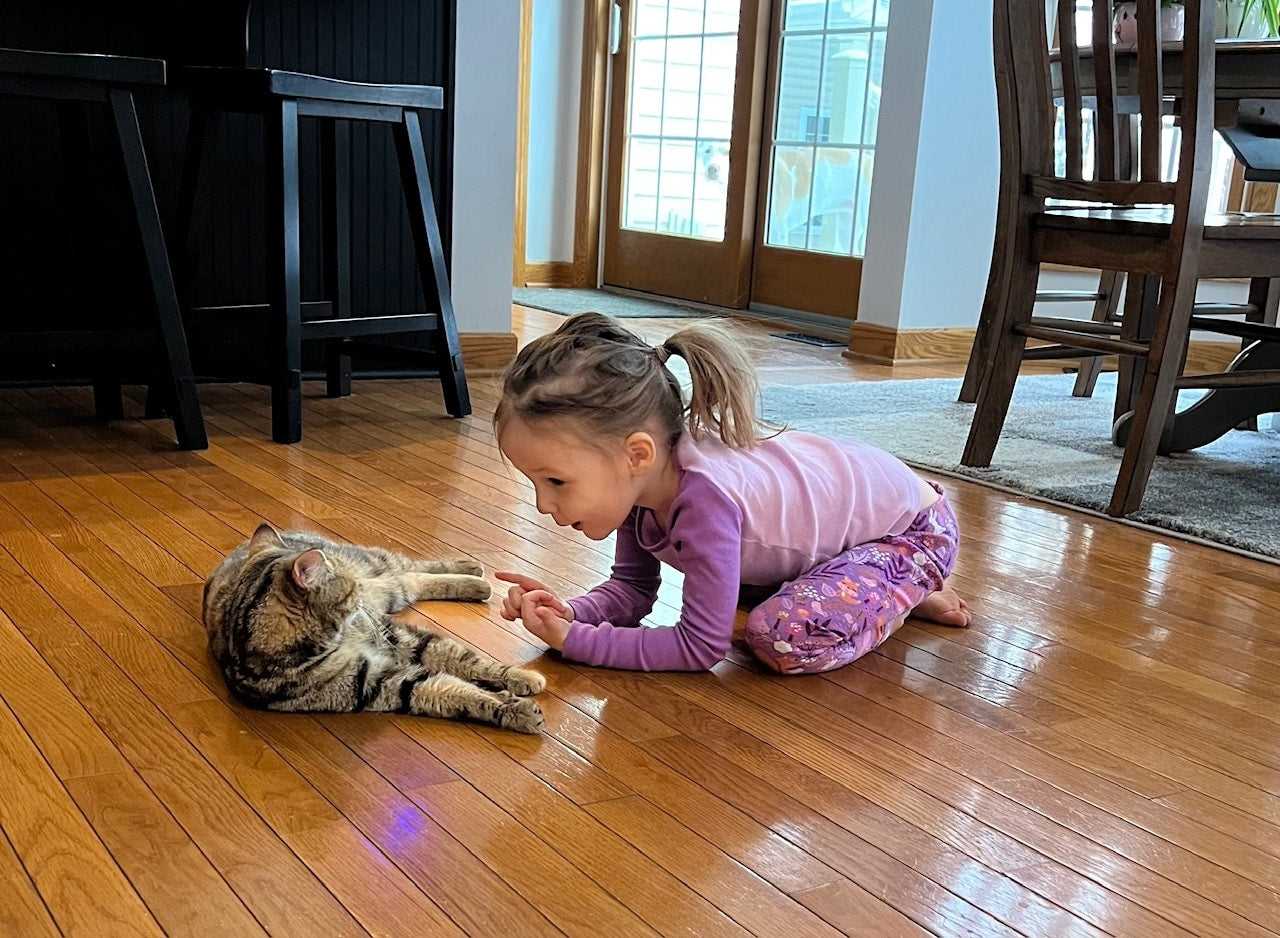 A young girl in purple pajamas gently engages with a tabby cat on a wooden floor in a cozy home setting. The scene conveys warmth and curiosity.