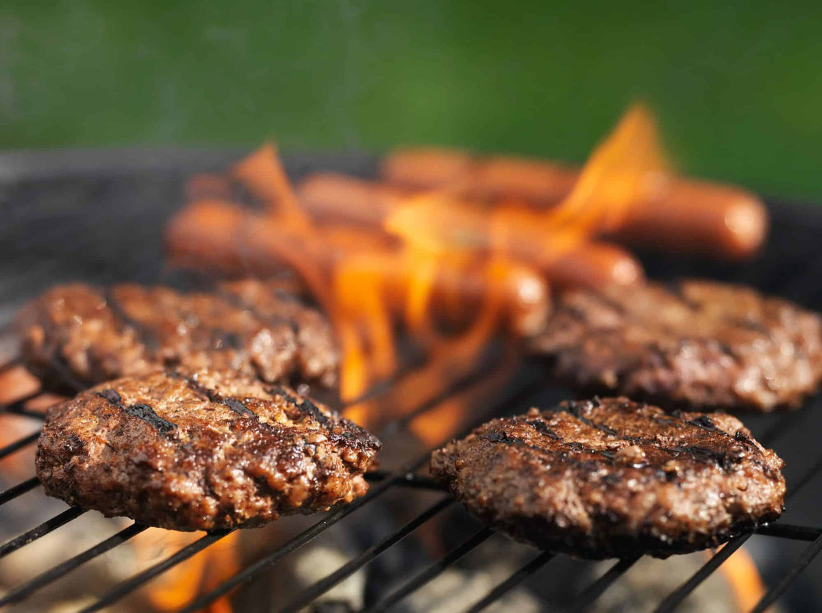 Close-up of a barbecue grill with sizzling burger patties and hot dogs over flames. The scene is lively, evoking a summer cookout.