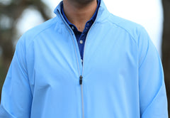 A person is wearing a light blue zip-up jacket over a dark blue shirt, with an out-of-focus natural background.