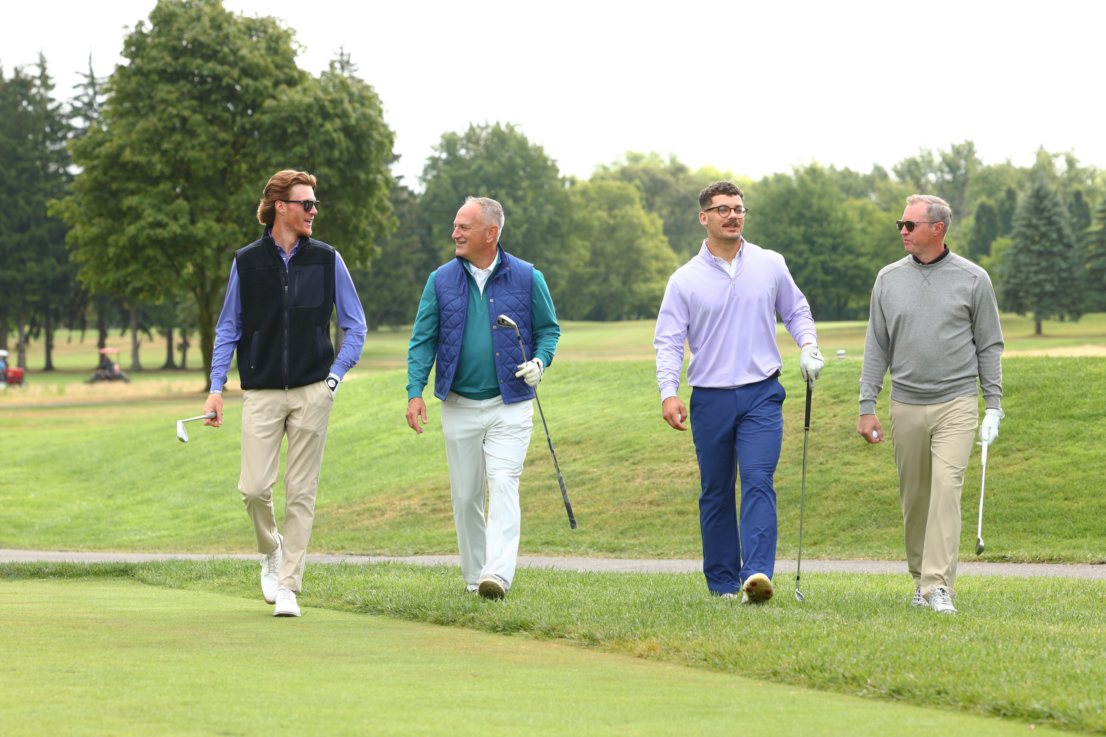 Four men walking on a golf course, each holding a club. They are casually dressed in sweaters and slacks, appearing relaxed and engaged in conversation.