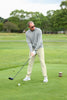 A man in a gray sweater and beige pants prepares to drive a golf ball on a lush green course. Trees are visible in the background under a cloudy sky.