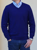 Man in a blue V-neck sweater over a checkered shirt, wearing jeans and a silver watch, standing against a plain beige background.