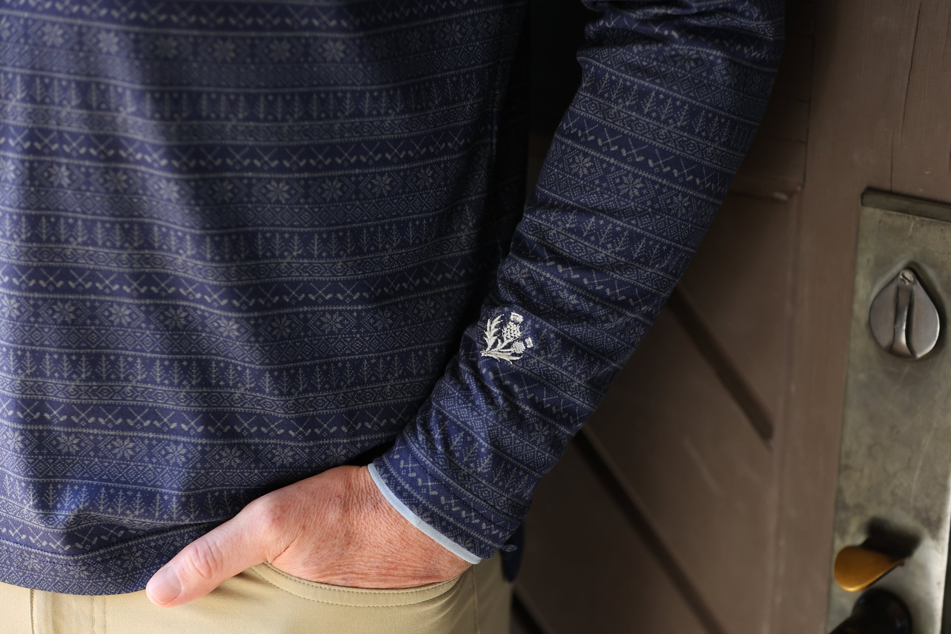 A person wearing a dark blue, patterned long-sleeve shirt stands near a wooden door. They have a hand in their tan pants pocket, creating a relaxed vibe.