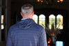 A person with short gray hair, wearing a patterned blue hoodie, stands facing a warmly-lit room with arched windows and decorative plants.