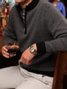 A golfer in a gray herringbone sweater and cream pants holds a drink in one hand and rests the other on a bar counter. They wear an elegant watch, conveying a relaxed, stylish vibe.