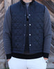 A person wearing a quilted navy vest over a patterned sweater is shown from the shoulders down. They have white pants and a silver watch, hands in pockets. The background features diagonal wooden planks.