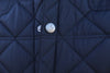 Close-up of a navy blue quilted jacket, featuring a prominent metal button engraved with a DJR logo and text. The fabric has a puffy diamond pattern.