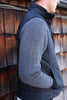 A person in a black quilted vest over a gray herringbone sweater stands sideways against a rustic wooden wall, conveying a cozy, autumnal feel.