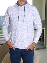 A man is standing with his hands in his pockets, wearing a white camo hoodie over a blue shirt and dark jeans.