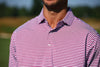Man wearing a magenta and white striped polo shirt standing outdoors with a blurred landscape background.