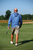 A golfer wearing a blue hoodie and khaki shorts walks across the fairway while holding a golf club.