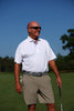 A man wearing a white polo shirt and khaki shorts stands on a golf course holding a putter under a clear blue sky.