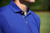 Man wearing a royal blue polo shirt with a patterned collar detail, photographed outdoors against a blurred green background.