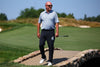 A golfer wearing a light blue patterned polo shirt and black trousers stands on a stone bridge crossing a golf course fairway on a sunny day.