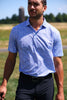 Man wearing a blue patterned polo shirt and dark trousers standing in an open field with trees and a silo in the background.