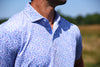 Man wearing a lavender and blue micro-floral print short-sleeve golf shirt with contrasting collar detail, photographed outdoors against a blurred natural background.