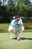 A man in a light blue polo shirt and cream pants lines up a putt on a golf green, wearing sunglasses and holding a putter.