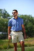 A man in a blue polo shirt and cream shorts walks across a golf course while holding a putter, wearing sunglasses on a clear day.