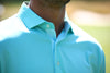 Man wearing a turquoise golf shirt with textured placket detail and light blue buttons, photographed outdoors on a course.