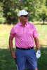 A golfer wearing a pink striped polo shirt and navy shorts stands on a manicured fairway while holding a golf club.