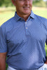 A man wearing a blue patterned short-sleeve polo shirt stands outdoors against a blurred green background.