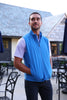 Man wearing a bright blue quilted vest over a light purple patterned short-sleeve shirt and dark trousers, standing on a patio outside a modern home.