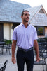 Man wearing a lavender short-sleeve polo shirt with navy trousers and a belt, standing on a patio in front of a modern home with gray shake roofing.