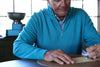 An older man in a bright blue quarter-zip sweater examines fabric details at a wooden table in a well-lit workspace.