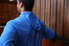 Man wearing a blue performance hoodie standing indoors with wood paneling in the background.