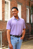 Man wearing a lavender polo shirt and blue jeans smiling in front of a brick building.