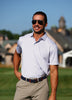 A man wearing a light blue patterned polo shirt and khaki trousers stands smiling on a golf course with a historic clubhouse visible in the background.