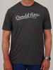 A man wearing a dark gray T-shirt with "Donald Ross Est. 1872" in white script. He has short sleeves, a watch, and blue pants, standing against a plain background.