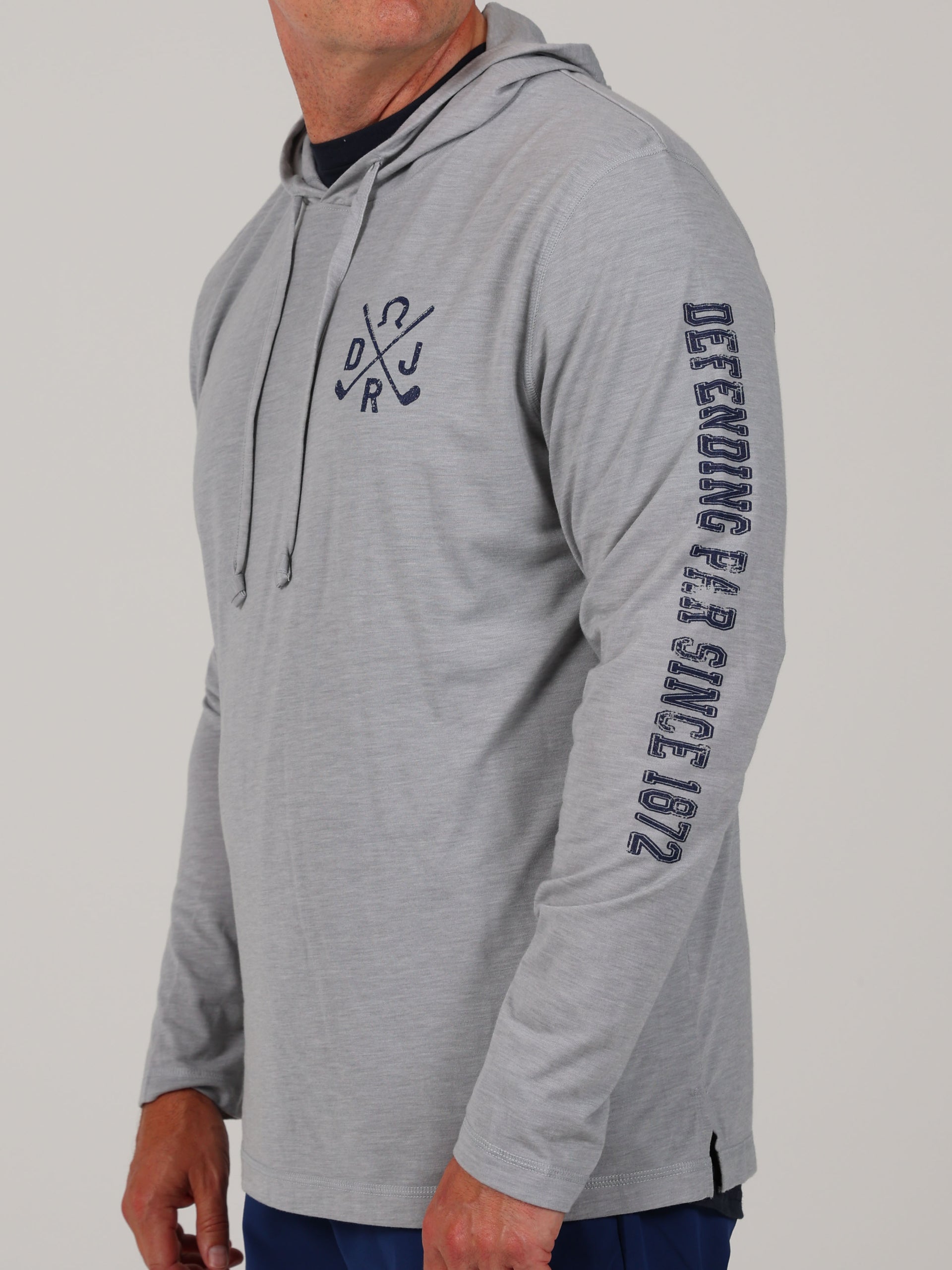 Man in a gray hooded sweatshirt with text on sleeve, "Defending Fair Since 1972," and crossed hockey sticks on chest. Casual, sporty vibe.