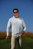 A man wearing a light gray long-sleeve hoodie and khaki trousers holds a golf club while standing on a golf course under clear blue skies.