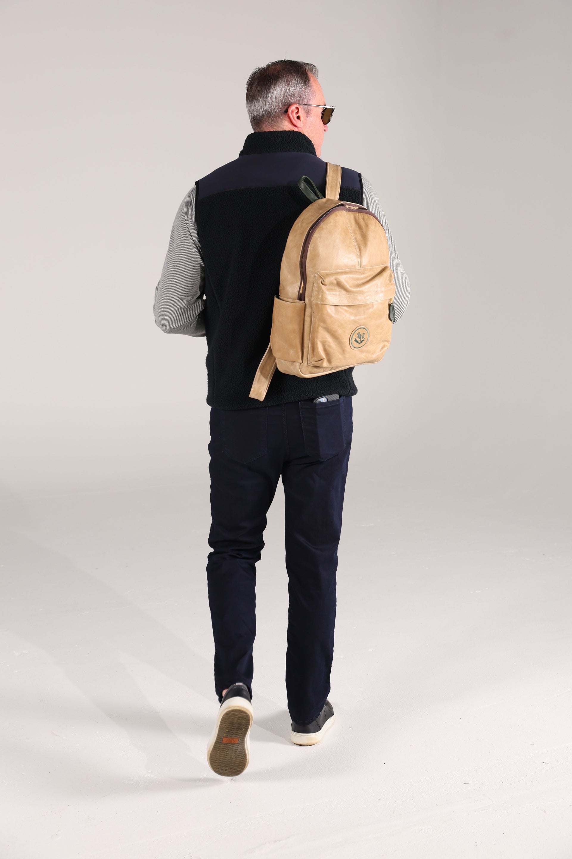 A man wearing a black vest, striped shirt, and dark pants walks away. He carries a tan backpack over one shoulder.